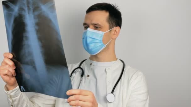 Egy fiatal sebész védőmaszkot visel, megvizsgálja a betegek tüdejének röntgenjét, és elgondolkodik a diagnózison.. — Stock videók