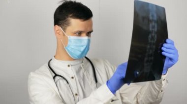 Tıbbi maskeli genç ve yakışıklı bir doktor ellerinde akciğerlerinin röntgen fotoğrafını tutuyor ve bir analiz yapıyor. Koruyucu maske takan genç bir cerrah hastaların röntgenlerini inceler.