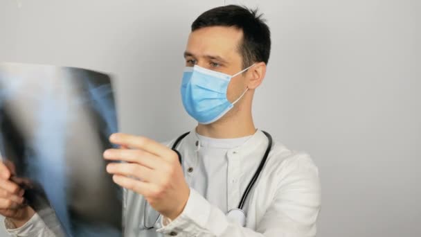 Młody przystojny lekarz w masce medycznej trzyma w rękach zdjęcie rentgenowskie płuc i przeprowadza analizę. — Wideo stockowe