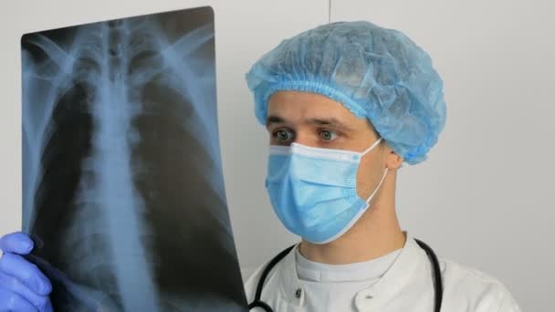 Un giovane chirurgo che indossa una maschera protettiva medica esamina una radiografia di un polmoni pazienti e medita la diagnostica.Un giovane bel medico in una maschera medica tiene una foto a raggi X dei polmoni nel suo — Video Stock