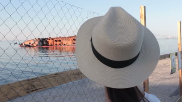 Beyaz şapkalı güzel bir kadın batık petrol tankerine bakıyor. Beyaz şapkalı genç çekici kadın, deniz kıyısında yatan batık tankere üzülerek bakıyor. Çevre felaketi. — Stok video
