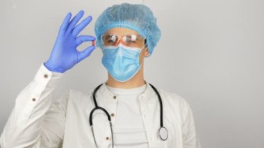 Tıbbi maskeli genç ve yakışıklı bir doktor elinde kırmızı bir hap tutuyor ve yeni bir ilaç sunuyor..