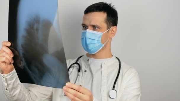 Un tânăr chirurg care poartă o mască medicală de protecție examinează o radiografie a plămânilor pacienților și analizează diagnosticul. . — Videoclip de stoc