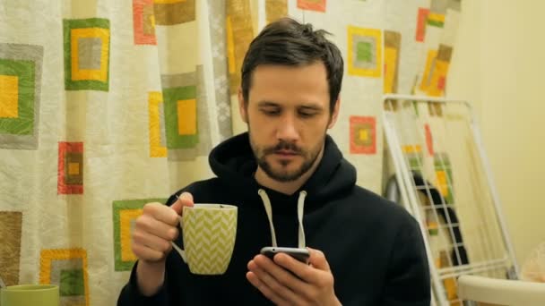 Joven chico atractivo está bebiendo café y volteando a través de las redes sociales de alimentación en su teléfono inteligente. Lindo chico joven está sentado en su sala de estar y beber café. — Vídeos de Stock