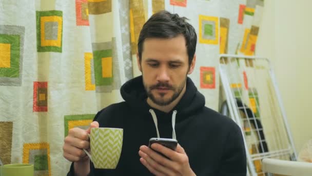 Egy vonzó fiatalember dolgozik az interneten az okostelefonján, miközben a konyhájában ül.Fiatal vonzó srác kávét iszik, és átlapozza a közösségi média feed az okostelefonján. Cuki. — Stock videók