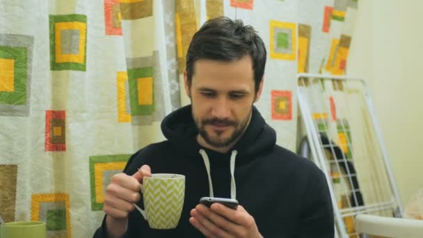 Joven chico atractivo está bebiendo café y volteando a través de las redes sociales de alimentación en su teléfono inteligente. Un joven atractivo está trabajando en Internet en su teléfono inteligente mientras está sentado en su cocina — Vídeos de Stock