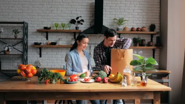 Um belo casal jovem de amantes de bom humor classificar legumes de um saco e colocá-los na mesa de cozinha.Jovem dona de casa feliz e jovem despreocupado colocar as compras em sua cozinha. Um jovem — Vídeo de Stock