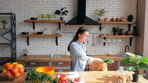 Happy young housewife lays out purchases in her kitchen.A beautiful young woman in a good mood is sorting vegetables from a bag and putting them on the kitchen table.Young Beautiful Woman lays out the — стоковое видео