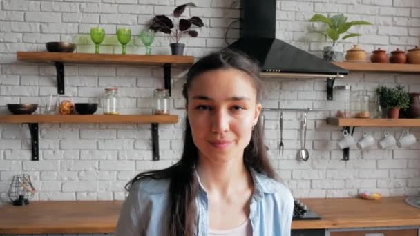 Feliz ama de casa joven mirando a la cámara, sonriendo ampliamente mientras está de pie en su cocina. retrato de cerca de una joven hermosa mujer que está mirando a la cámara y sonriendo ampliamente mientras — Vídeos de Stock