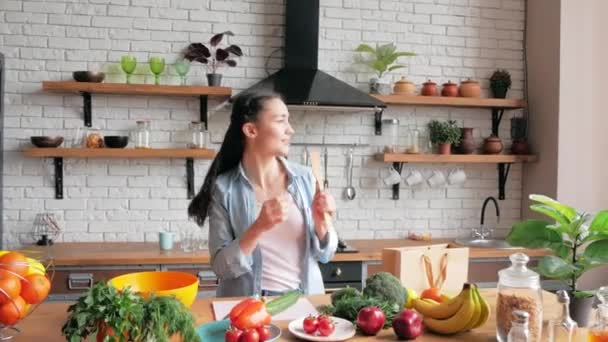 En glad ung hemmafru på gott humör sjunger roligt i en träsked och dansar glatt i sitt kök.En attraktiv ung kvinna sjunger och dansar medan hon lagar middag åt sin kärleksfulla pojkvän — Stockvideo