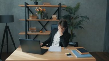 Genç ve yakışıklı bir iş adamı dizüstü bilgisayarına finansal bir rapor yazar ve yorgunluktan sandalyeye yaslanır. Genç ve çekici bir iş adamı uzun süre dizüstü bilgisayarda çalışır ve gözlerini ovuşturur.