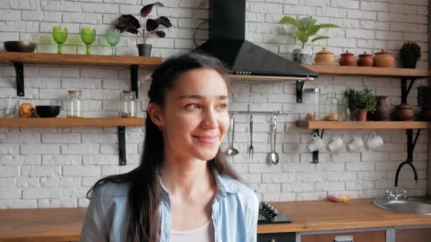 Feliz ama de casa joven mirando a la cámara, sonriendo ampliamente mientras está de pie en su cocina. De cerca retrato de una joven hermosa mujer que está mirando a la cámara y sonriendo ampliamente mientras — Vídeos de Stock