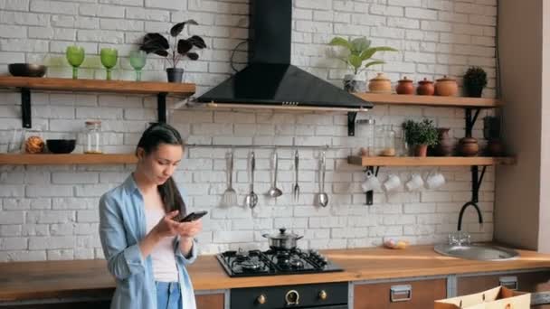 Jovem mulher bonita está virando através do feed de redes sociais em seu telefone enquanto está na cozinha. — Vídeo de Stock
