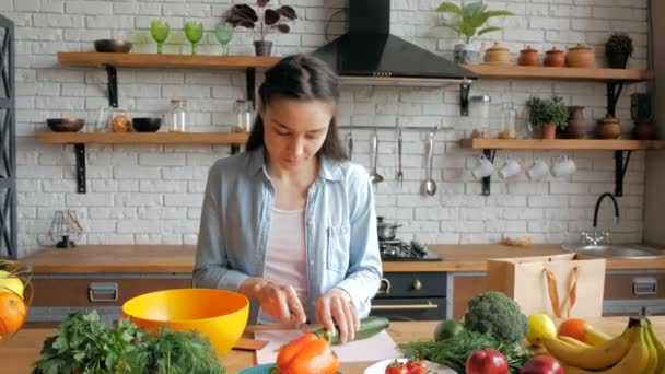 En vacker ung kvinna på gott humör skär en gurka för en sallad. Glad ung hemmafru tillagar grönsakssallad i köket. Ung vacker kvinna skär grönsaker för sallad och sätter dem i en — Stockvideo