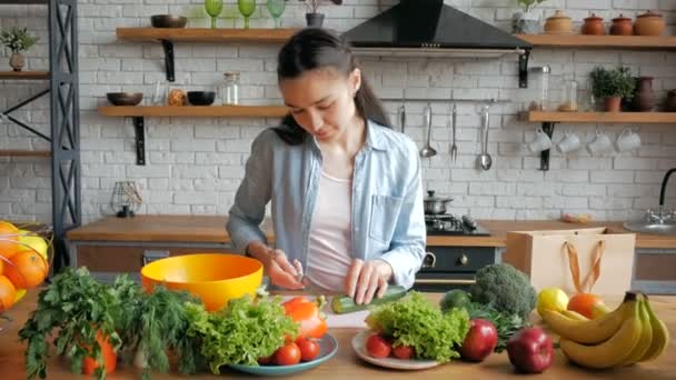 Eine schöne junge Frau schneidet gut gelaunt eine Gurke für einen Salat. Junge schöne Frau schneidet Gemüse für Salat und legt es in eine Schüssel. — Stockvideo