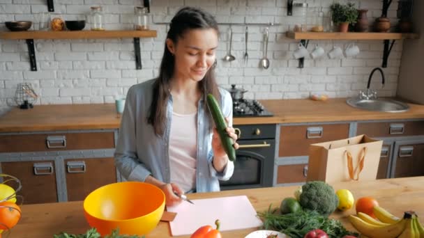 良い気分で美しい若い女性はサラダのためのキュウリをカットします。幸せな若い主婦は彼女の台所で野菜サラダを準備します。若い美しい女性はサラダのための野菜をカットし、それらを置く — ストック動画