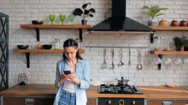 En glad ung hemmafru står i sitt kök och letar efter ett lämpligt recept på internet på sin smarta telefon. — Stockvideo
