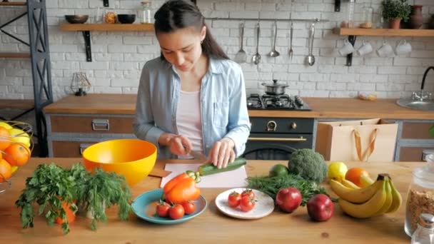 幸せな若い主婦は彼女の台所で野菜サラダを準備します。良い気分で美しい若い女性はサラダのためのキュウリをカットします。若い美しい女性はサラダのための野菜をカットし、それらを置く — ストック動画