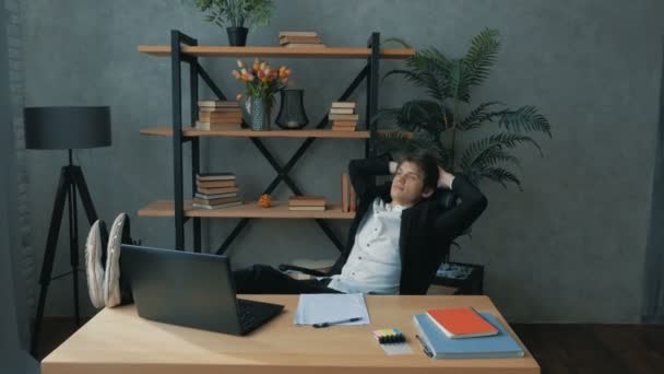 A young promising businessman drinks coffee carelessly with his legs on the table. Young handsome businessman writes a financial report on a laptop and leans back in a chair while resting from work — Stock Video