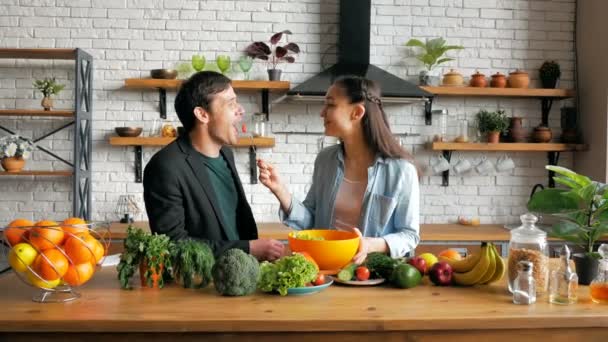 Una joven ama de casa feliz invita a su marido a cenar en su cocina. Joven hermosa mujer junto con su amado marido preparar una ensalada de verduras en la cocina de su apartamento. — Vídeos de Stock