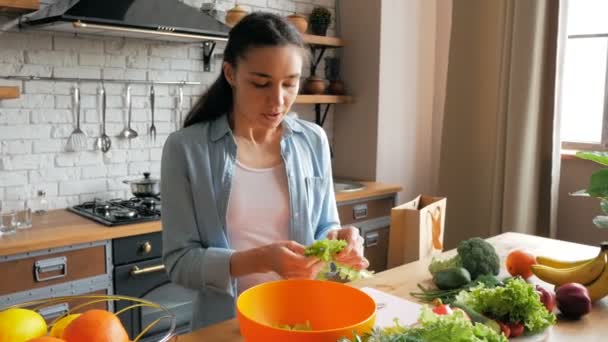 漂亮的年轻女子心情很好,为沙拉切莴苣叶.年轻漂亮的女人切蔬菜当沙拉,放在一个碗里.快乐的年轻家庭主妇在自己家里准备蔬菜沙拉 — 图库视频影像