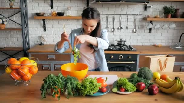 En vacker ung kvinna på gott humör rör om i den kokta salladen i en apelsinskål. Glad ung hemmafru tillagar grönsakssallad i köket. Ung vacker kvinna skär grönsaker för sallad — Stockvideo