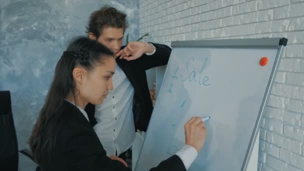 Молодой парень и красивая привлекательная девушка обсуждают рост продаж, рисуя график на черной доске. Офисные работники в деловых костюмах обсуждают концепцию нового ИТ-продукта, рисуют — стоковое видео
