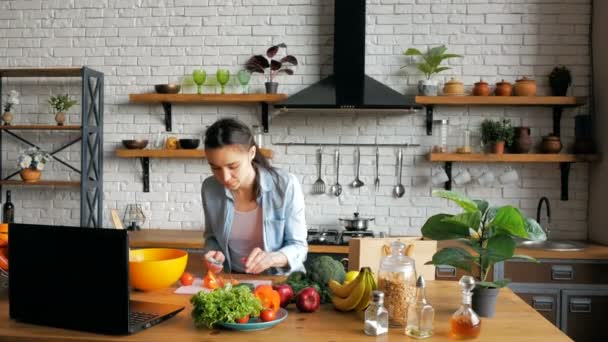 Gute gelaunte junge Hausfrau bereitet nach einer Videochat-Stunde einen Salat zu. Glückliche junge Hausfrau bereitet Gemüsesalat in der Küche zu, während sie sich ein Rezept im Internet auf ihrer — Stockvideo