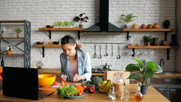 Feliz ama de casa joven de buen humor prepara una ensalada después de una lección de video chat. Feliz joven ama de casa preparando ensalada de verduras en la cocina mientras mira una receta en Internet en su — Vídeo de stock