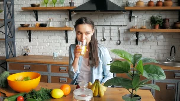 Seorang wanita muda yang menarik adalah minum jus jeruk di dapur dan menunggu pacar yang dicintainya. Happy young housewife in a good mood is joined freshly squeezed orange juice while sitting — Stok Video