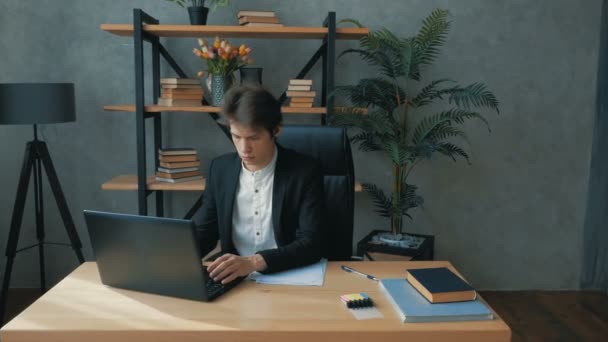 A young attractive businessman works on a laptop for a long time and wearily rubs his eyes while sitting at a table in his office. A young handsome businessman writes a financial report on a laptop — Stock Video