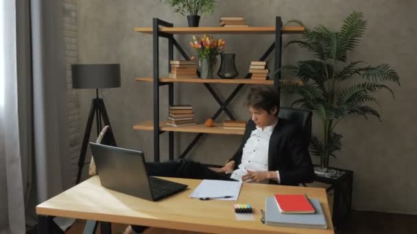 Young attractive businessman is resting from work with his legs on the table. Young handsome businessman writes a financial report on a laptop and leans back in a chair while resting from work. — Stock Video