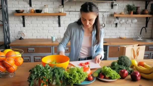 Vacker ung kvinna på gott humör skär mogen körsbärstomat för sallad. Ung vacker kvinna skär grönsaker för sallad och lägger dem i en skål. Lycklig ung hemmafru förbereder grönsakssallad i sin — Stockvideo