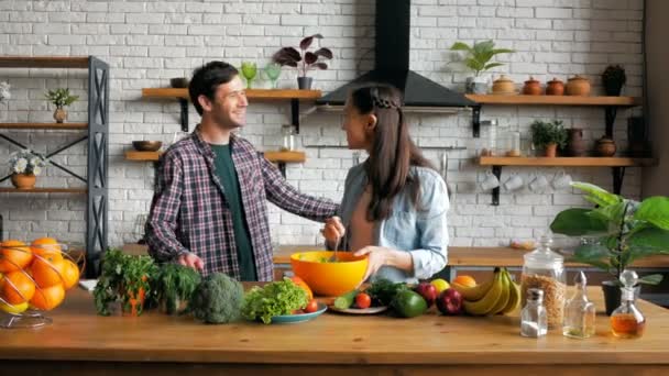 Una joven ama de casa feliz invita a su marido a cenar en su cocina. Joven hermosa mujer junto con su amado marido preparar una ensalada de verduras en la cocina de su apartamento. — Vídeo de stock