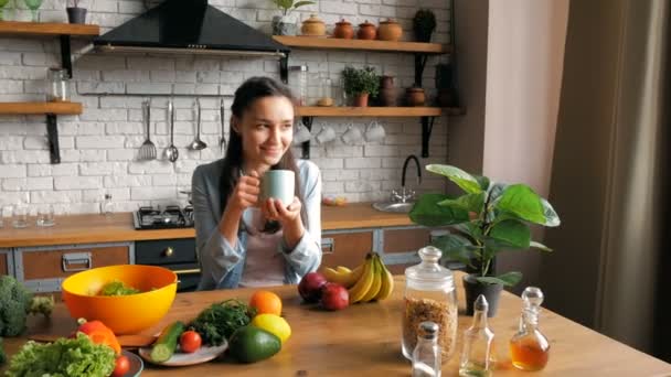 O tânără atractivă bea cafea aromată în bucătărie și își așteaptă iubitul. O gospodină tânără fericită într-o stare bună se bucură de cafea proaspăt preparată în timp ce stă la o masă în — Videoclip de stoc