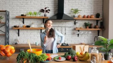 Neşeli genç bir ev hanımı, neşeli bir ruh hali içinde, metal bir kaşıkla komik bir şekilde şarkı söyler ve mutfakta neşeyle dans eder. Genç kaygısız kadın mutfakta dans eder ve şarkı söyler..