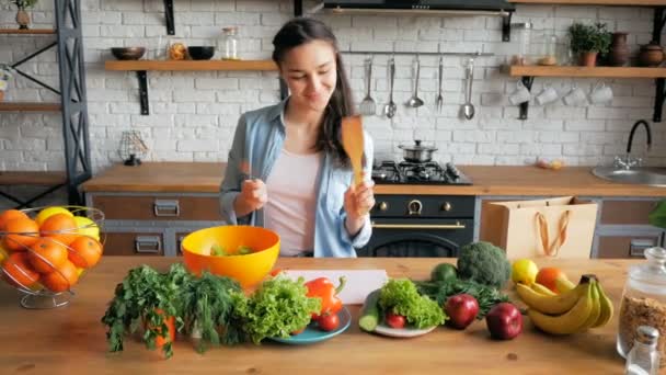 A beautiful young woman in a good mood is stirring the cooked salad in an orange bowl. Happy young housewife prepares vegetable salad in her kitchen. Young beautiful woman cuts vegetables for salad — Stock Video