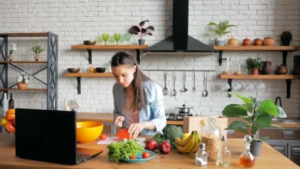 Una joven hermosa mujer, siguiendo una receta en Internet, corta verduras para una ensalada y las pone en un boliche. La joven ama de casa feliz de buen humor prepara una ensalada después de una lección de video chat — Vídeo de stock