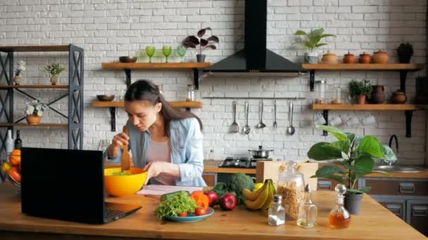 Felice giovane casalinga preparare insalata di verdure in cucina mentre guardando una ricetta su internet sul suo computer. Felice giovane casalinga di buon umore prepara un'insalata dopo una video chat — Video Stock