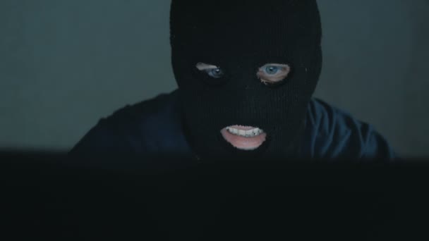 Un hacker de Internet en un pasamontañas negro se sienta en una silla y roba datos de una computadora portátil. Un hacker anónimo con capucha negra llama a su víctima por teléfono y amenaza con descifrar las contraseñas de la base de datos — Vídeos de Stock