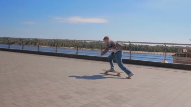 Jovem atraente está montando um skate. Um skatista profissional monta um skate ao longo do dique da cidade. Um jovem milenar que monta um skate despreocupado na cidade. — Vídeo de Stock