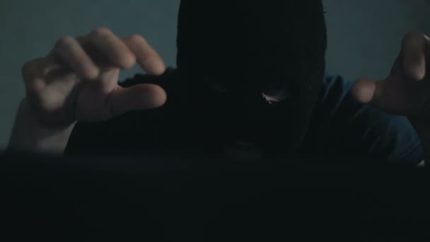一个穿着黑色巴拉克拉瓦的网络黑客坐在椅子上，从笔记本电脑上窃取数据。一名身穿黑色帽衫的匿名黑客打电话给受害者，威胁要破解数据库密码 — 图库视频影像