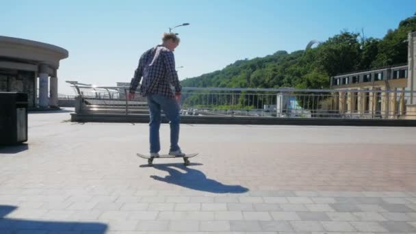 A professional skateboarder rides a skateboard along the city embankment. A young millennial guy who rides a skateboard carefree in the city. The professional skateboarder performs tricks to improve — Wideo stockowe