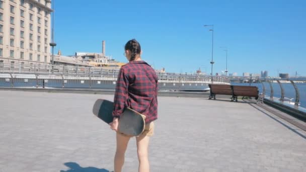 Junge Skateboarderin, die mit einem Skateboard die Straße entlangläuft, dreht sich um und lächelt in die Kamera. Junge attraktive Millennial-Frau läuft die Fußgängerpromenade entlang — Stockvideo