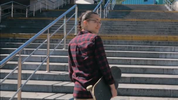 Mladá atraktivní tisíciletá žena kráčí po schodech se skateboardem v rukou. Mladá žena skateboardista stoupá po schodech otočí a usmívá se při pohledu do kamery. Zavřít portrét — Stock video