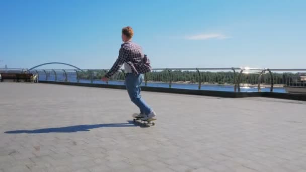 Jovem atraente está montando um skate. Um skatista profissional monta um skate ao longo do dique da cidade. Um jovem milenar que monta um skate despreocupado na cidade. O — Vídeo de Stock