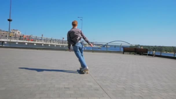 En professionel skateboarder rider et skateboard langs byens dæmning. Ung attraktiv fyr kører på skateboard. En ung tusindårig fyr, der rider et skateboard sorgløs i byen. Den – Stock-video