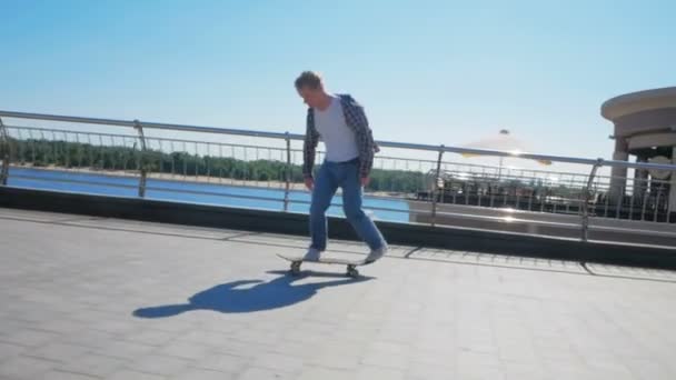 Jovem atraente está montando um skate. Um skatista profissional monta um skate ao longo do dique da cidade. Um jovem milenar que monta um skate despreocupado na cidade. O — Vídeo de Stock