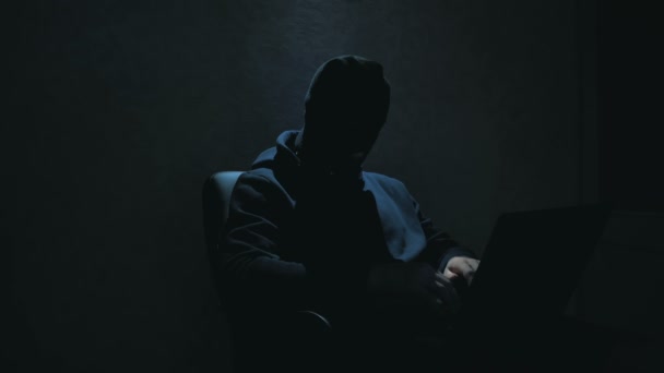 Ein Cyber-Terrorist mit schwarzer Sturmhaube hackt sich in eine Webseite und stiehlt Internetnutzern wichtige Daten. Ein Internet-Hacker mit schwarzer Sturmhaube sitzt auf einem Stuhl und stiehlt Daten von einem Laptop — Stockvideo