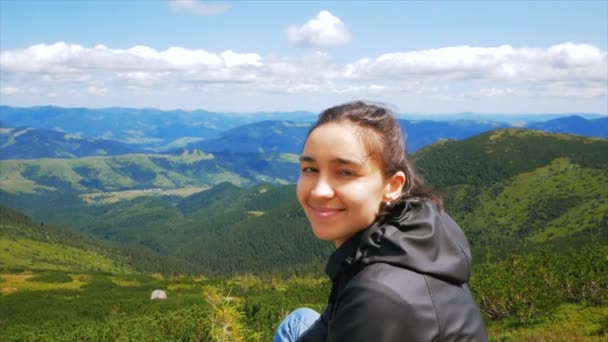 若いハイカーの女性と笑みを浮かべて、カメラの崖の上の山の風景の背景に座って見て。若い女性旅行者が山の頂上に到達し、美しいを楽しんでいます — ストック動画